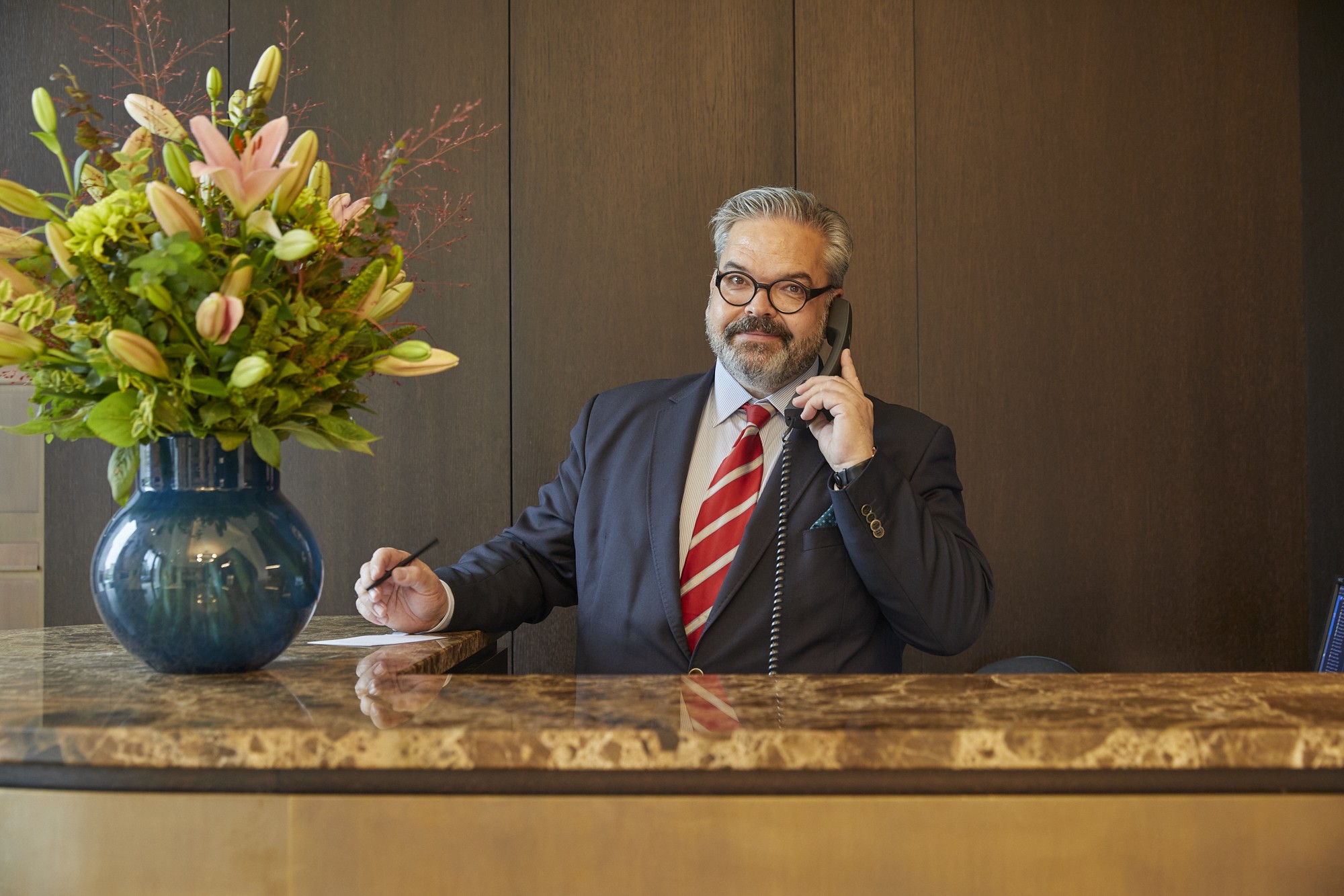 Concierge am Empfangstresen aus Marmor beim Telefonieren, daneben eine Vase mit Lilien