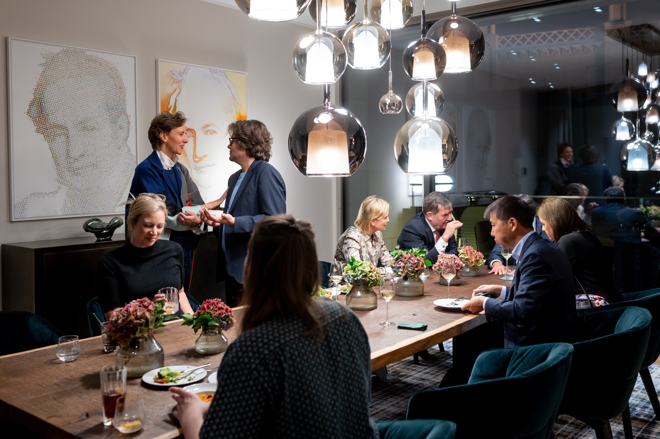 Gäste an einer langen Tafel unter gläsernen Pendelleuchten bei einem Abendessen in der Eventlocation