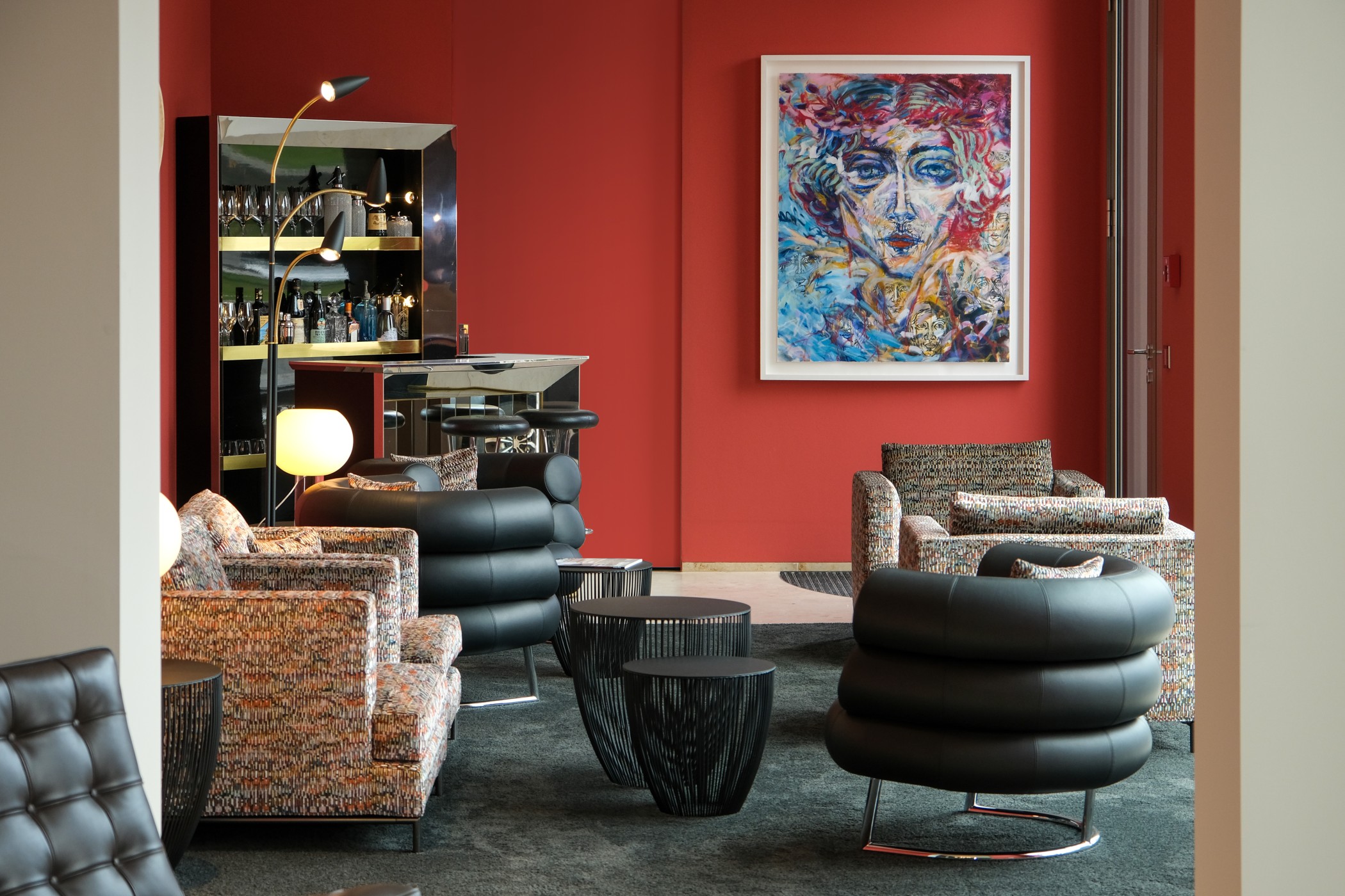 Lounge mit roter Wand, farbigem Porträtgemälde, Bar, gemusterten Sesseln und schwarzen Polstersesseln