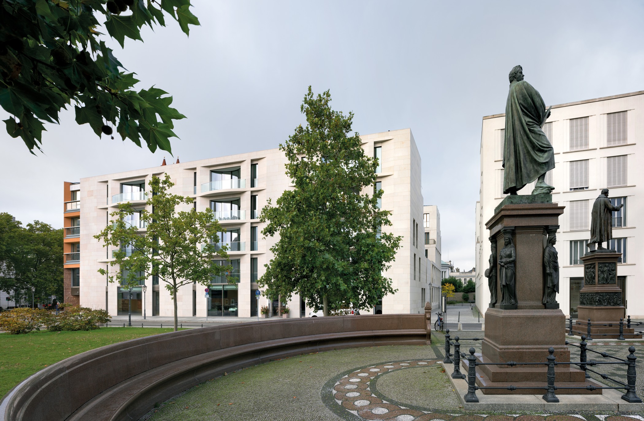 Zwei Denkmäler am Schinkelplatz mit Rasenfläche und geschwungener Steinbank vor einem hellen Wohnhaus