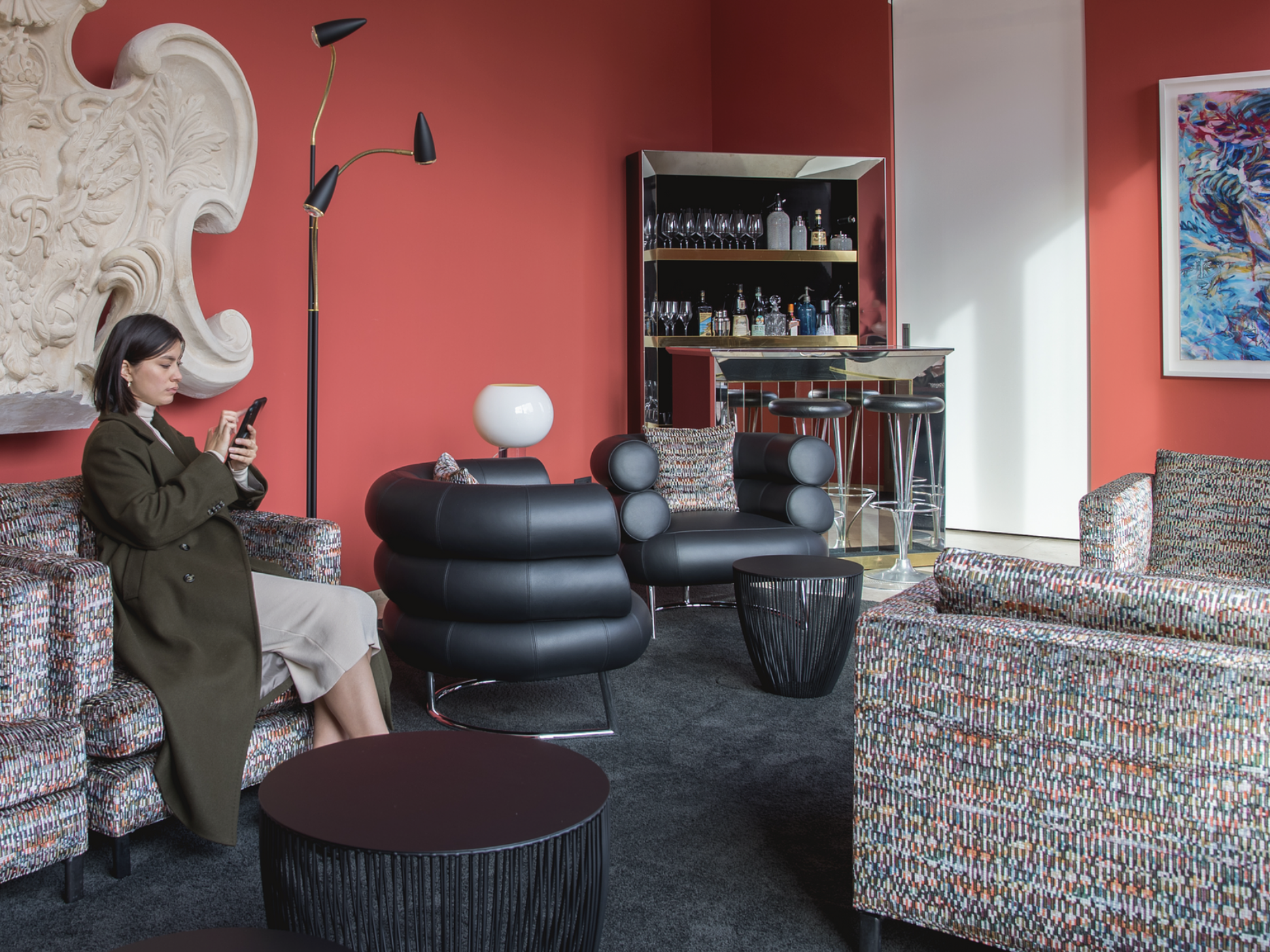 Frau mit Smartphone in einem gemusterten Sessel der Lounge, daneben schwarze Polstersessel und Bar