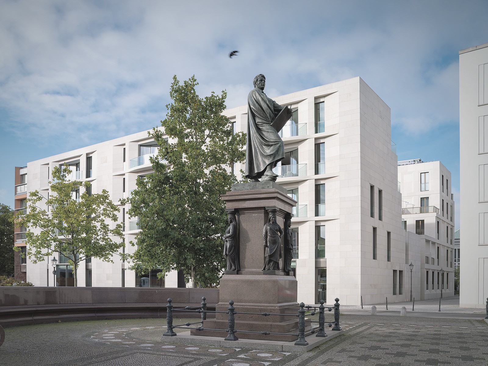 Schinkel-Denkmal mit eiserner Umzäunung vor hellen Wohngebäuden und Bäumen am Schinkelplatz