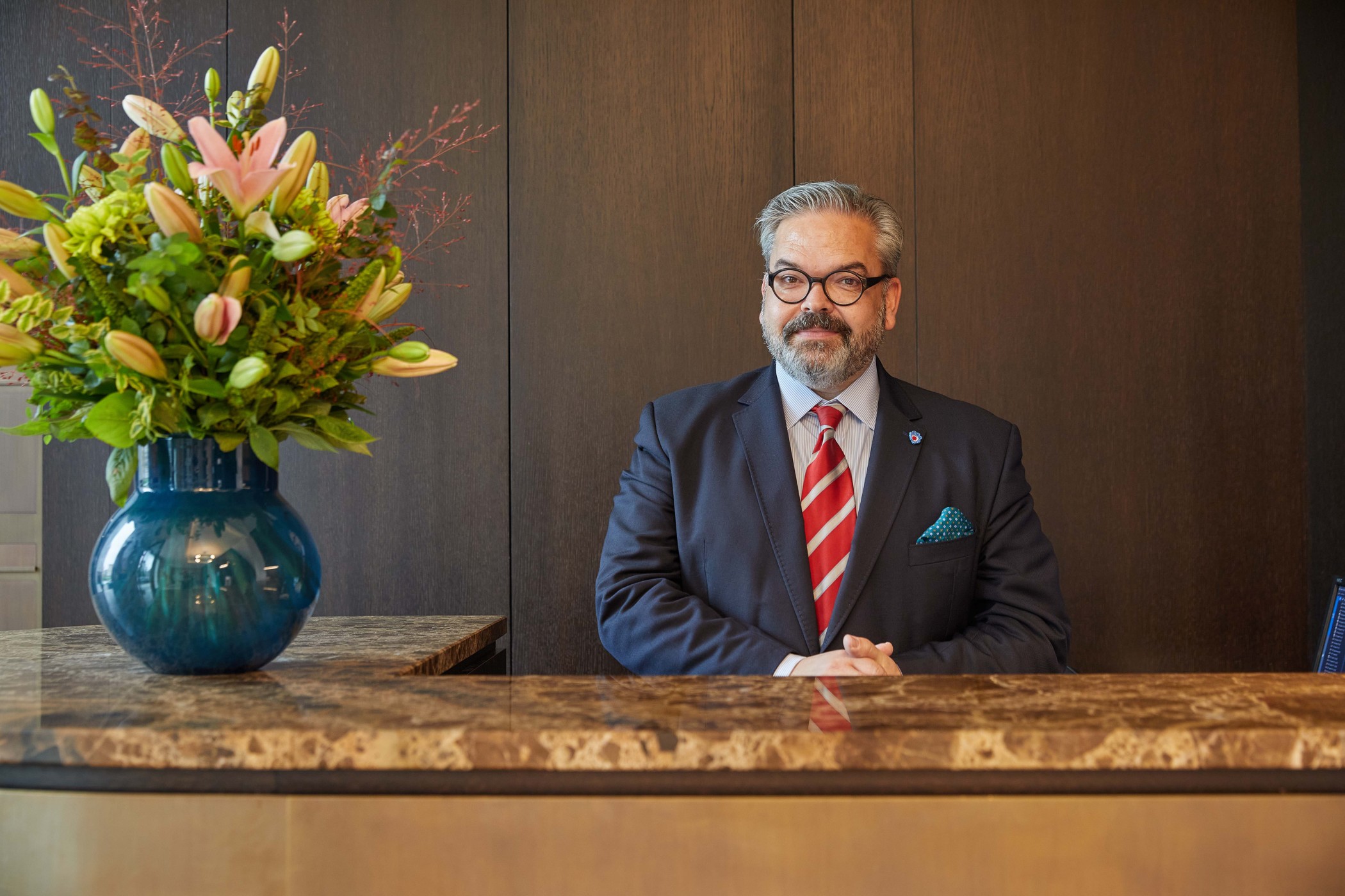 Concierge mit gefalteten Händen hinter dem Empfangstresen aus Marmor, daneben eine Vase mit Lilien