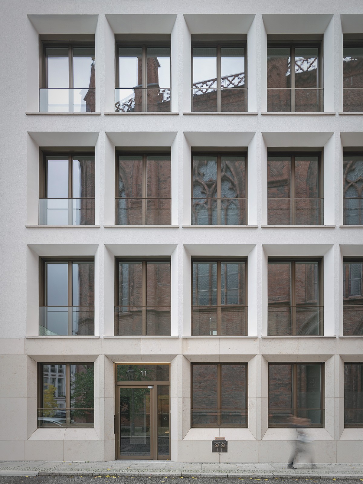 Rasterfassade aus hellem Naturstein mit tiefen Fensterlaibungen, in denen sich eine Backsteinkirche spiegelt
