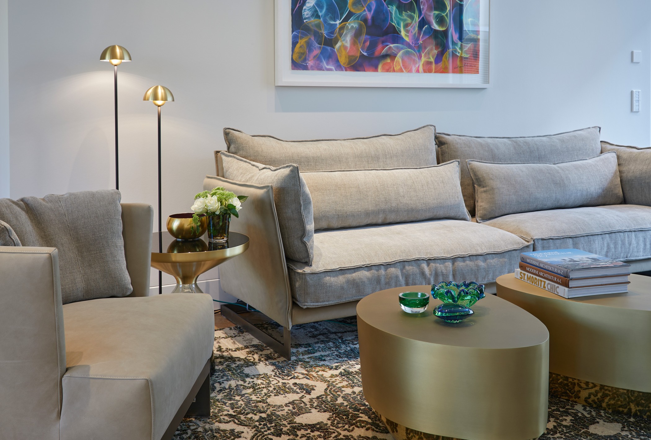 Wohnzimmer mit beigefarbenem Sofa, goldfarbenen runden Couchtischen, zwei Stehleuchten und farbenfrohem Gemälde