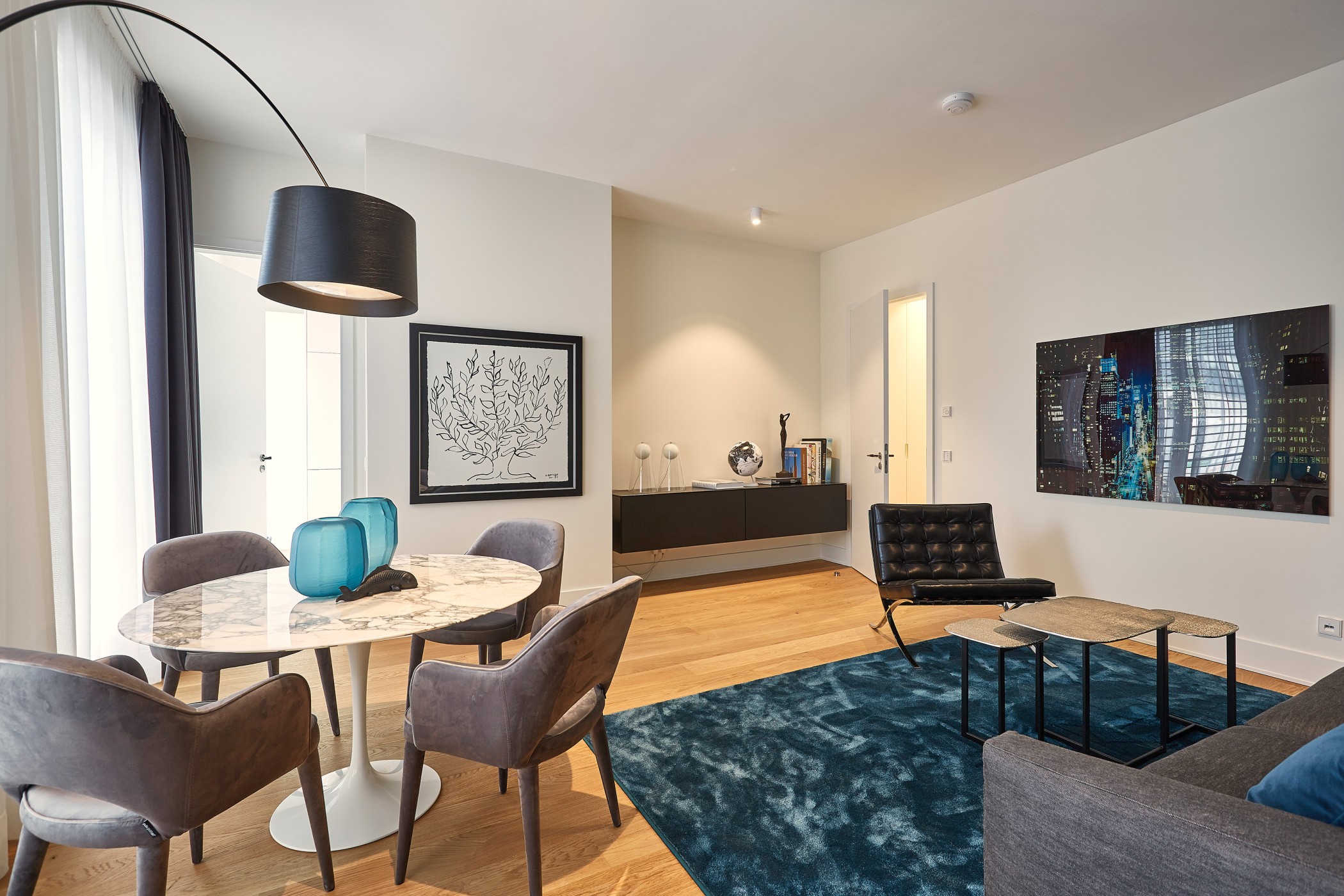 Wohn- und Essbereich mit rundem Marmortisch, blauen Glasvasen, Bogenleuchte, blauem Teppich und Kunstwerken an den Wänden