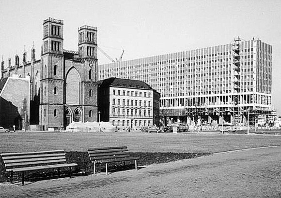 Historische Schwarzweißaufnahme der Friedrichswerderschen Kirche neben einem im Bau befindlichen Großbau, davor Rasen mit Bänken