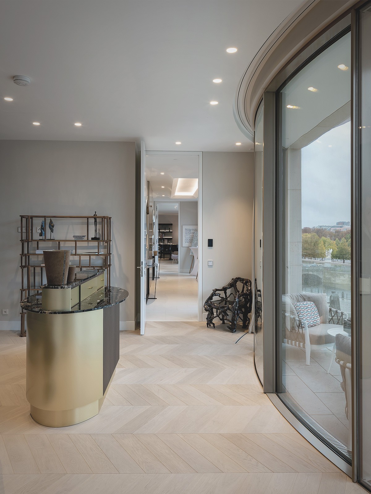 Flur einer Wohnung mit geschwungener Glasfront, Fischgrätparkett und goldfarbener Konsole mit Dekoobjekten