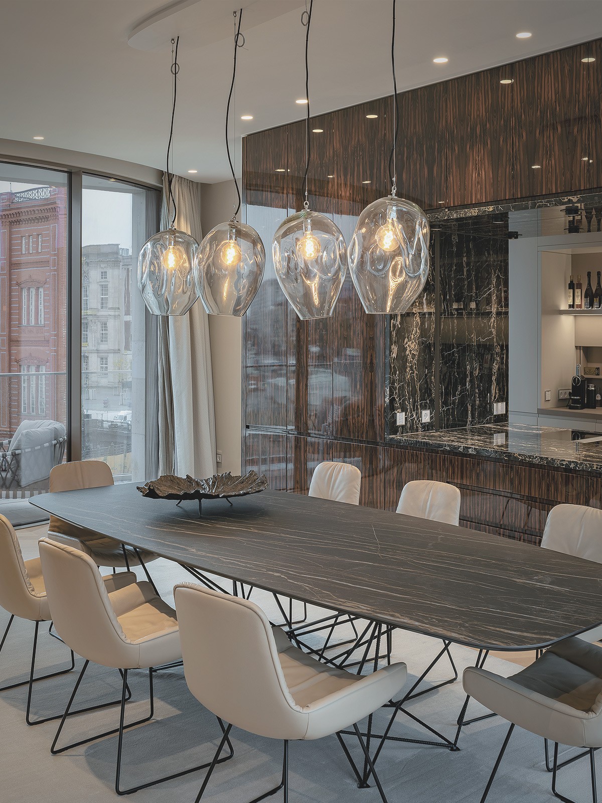 Essbereich mit langem Tisch, hellen Stühlen und gläsernen Pendelleuchten vor einer Wand aus dunklem Holz und Marmor